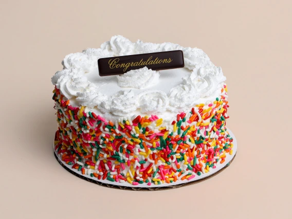 A round vanilla cake covered in white frosting, decorated with colorful rainbow sprinkles on the sides, and topped with piped whipped cream swirls. A small chocolate plaque with the word 'Congratulations' in gold script rests on top. The cake sits on a white cake board against a neutral beige background.